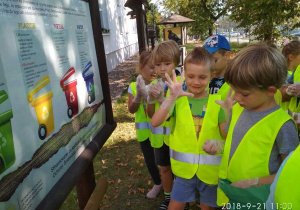 Dzieci w odblaskowych kamizelkach oraz rękawiczkach przy tablicy dydaktycznej dotyczącej segregacji odpadów