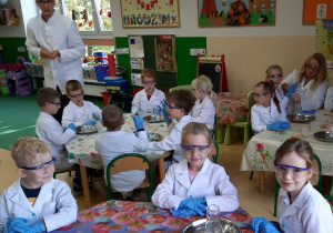 Dzieci podczas warsztatów, siedzą przy stolikach, na stołach stoją tace ze szklanymi butelkami