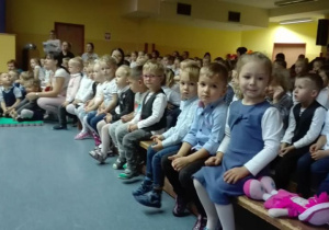 Dzieci podczas uroczystości, sidzą na ławeczkach na auli