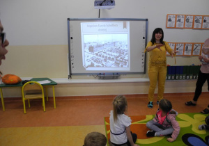Dzieci podczas zajęć, słuchają Pani, która pokazuje na tablicy multimedialnej zdjęcie łódzkiej fabryki z dawnych lat