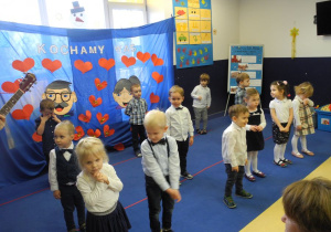 Dzieci podczas uroczystości, stoją na scenie, za nimi napis Kochamy Was i rysunek głowy babci i dziadka oraz serca