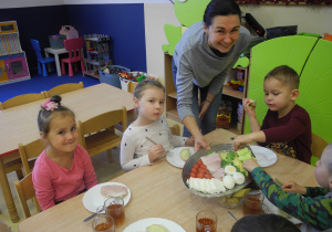 Dzieci przygotowują i jedzą kanapki, pani dietetyk częstuje je produktami z półmiska. Dwóch chłopców bierze z półmiska ser żółty. Na stoliku stoją szklanki z herbatą oraz bułki posmarowane masłem