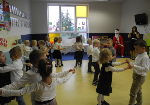 Dzieci ustawione w parach naprzeciwko siebie po kole. W tle widzimy okno, a na nim ozdoby świąteczne