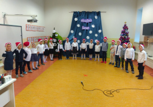 Dzieci z grupy Sowy ubrane na galowo, stoją w półkolu w czapkach mikołajowych