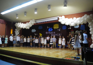 Zdjęcie grupowe Kotów na scenie, w tle napis witajcie wakacje na granatowym tle, dookoła napisu chmurki oraz słoneczko