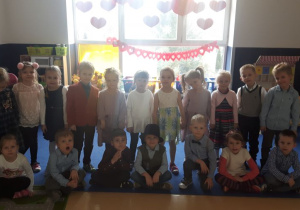 Zdjęcie grupowe Kangurków na tle okna, na oknie naklejone czerwone serca z tektury i girlanda z serc
