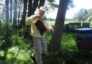 Pan Pszczelarz w wstoju pszczeraskim trzyma w ręku ramkę z miodem. Stoi wśród drzew, po prawej stronie stoją ule