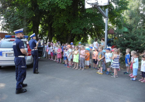Policjant i Policjantka stoją obok radiowozu. Naprzeciwko nich stoją dzieci. Słuchają wypowiedzi na temat pracy w policji
