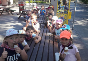 Dzieci siedzą przy drewnianej ławie, jedzą lody