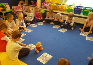 Dzieci podczas warsztatów, siedzą na dywanie, mają przed sobą kartkę z historyjką obrazkową