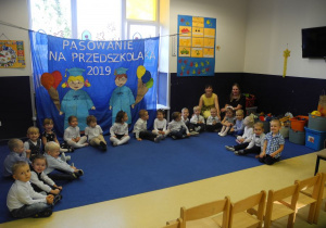 Dzieci przed uroczystością, siedzą wraz z Paniami na dywanie, w tle napis: Pasowanie na przedszkolaka 2019 oraz postać chłopca i dziewczynki
