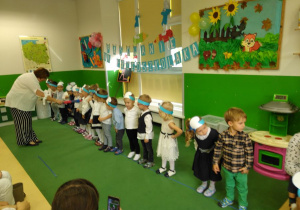 Pani Dyrektor przyjmuje dzieci w poczet Przedszkolaków, dotyka ołówkiem ramienia chłopca, dzieci stoją w szeregu czekając na swoją kolej