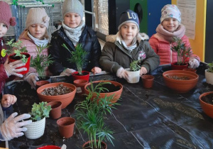Dzieci na warsztatach ogrodniczych, szykują się do sadzenia kwiatów, stoją przy stole, na którym stoją doniczki z ziemią i doniczki z kwiatami
