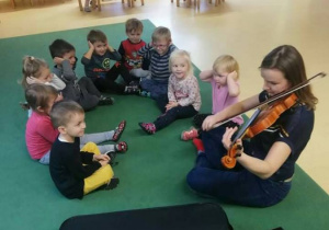 Dzieci podczas zajęć muzycznych, siedza z pania na dywnaie, Pani gra na skrzypcach