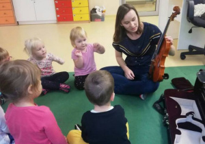 Dzieci podczas zajęć muzycznych, słuchają Pani, która pokazuje skrzypce i opowiada o nich dzieciom