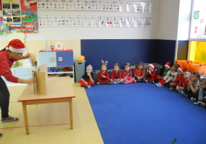 Dzieci siedzą w półkolu słuchają opowiadania czytanego przez nauczycielkę, która korzysta z teatrzyku Kamishibai, w tle alfabet