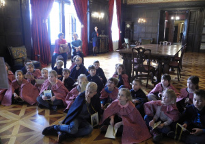 Dzieci siedzą w sali pałacowej w Pałacu Herbsta, słuchają oprowadzającej