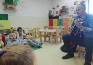 Dzieci siedzą na dywanie i słuchają Pana, który gra na gitarze